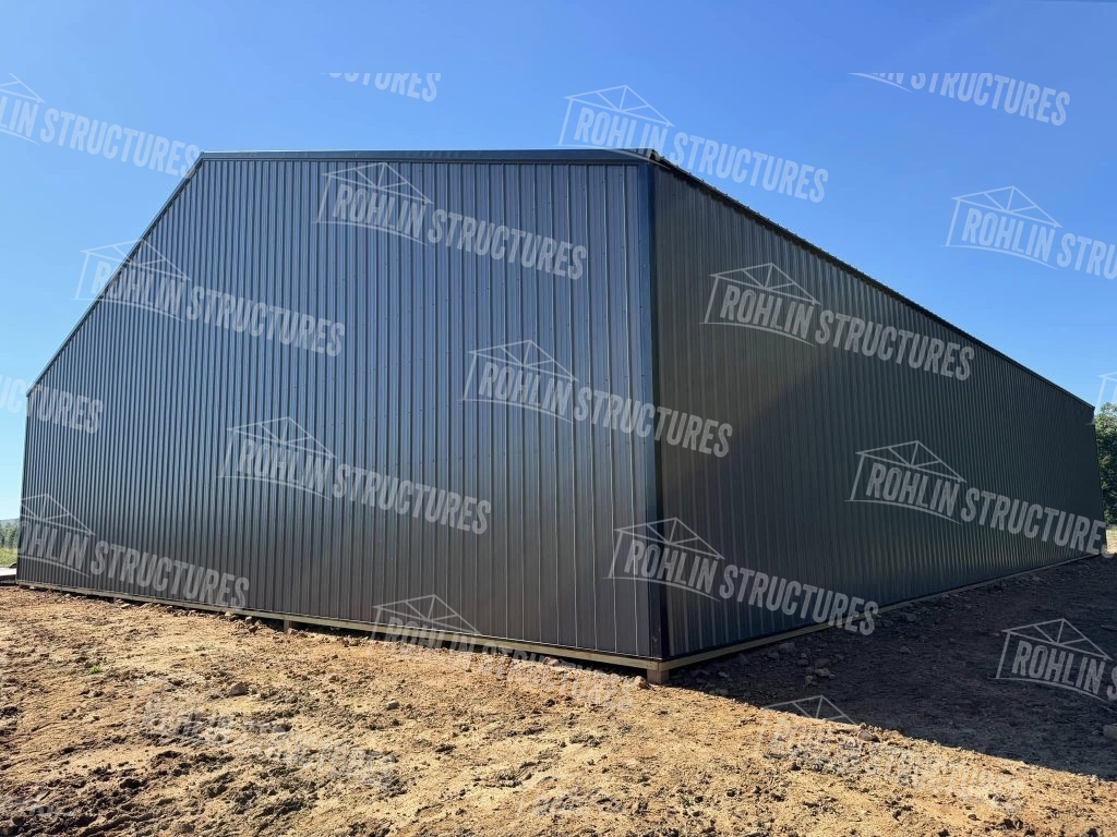 A large, dark gray Rohlin Structures equipment cover building stands on a dirt lot.